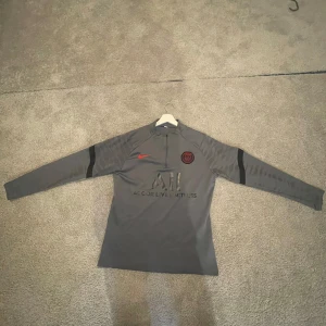 Grå långärmad PSG Nike fotbollströja - Säljer en grå långärmad PSG-tröja från Nike med svart PSG-logga och röd Nike-swoosh på bröstet. Tröjan har halv dragkedja, svarta detaljer på ärmarna och tryck med 'Accor Live Limitless' och 'Visit Rwanda'. Materialet är lätt och andas, perfekt för träning.