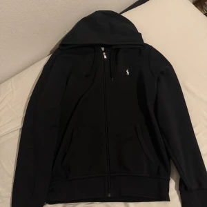 Svart hoodie från Ralph Lauren - Svart hoodie från Ralph Lauren med dragkedja och klassisk logga på bröstet. Tröjan har huva med snörning och två stora fickor framtill. Perfekt för en chill och stilren look. Ena snöret gick sönder i tvätten 
