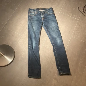 Blå Nudie Jeans, 28/32 - Säljer ett par klassiska blå jeans från Nudie Jeans i storlek 28/32. Jeansen har raka ben, snygga slitningar och kontrastsömmar. Tillverkade i bomull med femficksdesign och detaljerade bakfickor. Perfekta för dig som gillar en tidlös och clean look.