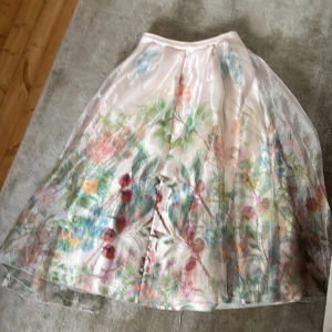 Blommig långkjol från H&M - Superfin långkjol från H&M med ett skirt lager och blommigt mönster i pastellfärger. Kjolen har en ljusrosa bas och är gjord i polyester med ett glansigt yttre. Perfekt för dig som vill ha en romantisk och drömmig look.