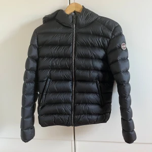 Svart vinterjacka från Colmar med huva - Snygg svart vinterjacka från Colmar med huva och dragkedja framtill. Jackan har en quiltad design, två fickor med dragkedja och en diskret logga på ärmen. Perfekt för kalla dagar när du vill hålla stilen och värmen. Italiensk storlek 46, motsvarar 38-40 i EU