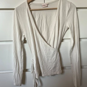 Vit omlottopp från Nelly - Säljer en stilren vit omlottopp från Nelly i mjuk bomull. Toppen har långa ärmar, djup v-ringning och knyt i sidan för en snygg passform. Perfekt att styla med jeans eller kjol för en clean look.