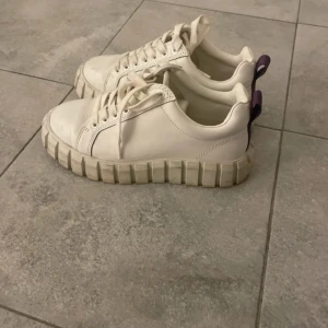 Chunky vita sneakers från Eytys - Säljer ett par chunky vita sneakers från Eytys med kraftig platt sula och lila hälflik. Skorna har snörning, rund tå och är tillverkade i skinn. I storlek 38 och knappt använda. Har lådan kvar från eytys. Ordinarie pris 2200kr