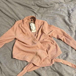 Ribbad omlottopp i blush från Boohoo - Supersnygg ribbad omlottopp i blushrosa från Boohoo. Toppen har långa ärmar, knytband i midjan och en v-ringad omlottfront. Perfekt för dig som gillar en feminin och trendig look med extra detalj i knytet framtill.