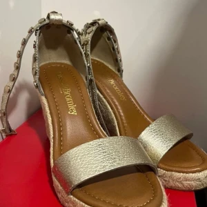 Guldiga wedge-sandaler Russell & Bromley - Snygga sandaler i guldigt skinn från Russell & Bromley med kilklack i rep och coola nitar längs remmarna. Öppen tå och justerbar ankelrem. Perfekta för sommaren när du vill sticka ut lite extra.