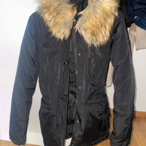 Svart parkajacka med fuskpäls Roco Baroco - Svart parkajacka från Roco Baroco med stor, fluffig fuskpäls i beige och guldtoner runt kragen. Jackan har dragkedja och knappar framtill, flera fickor och markerad midja. Perfekt för kalla dagar och riktigt snygg till vintern.