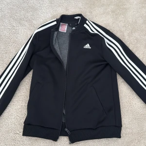 Svart Adidas bomberjacka med vita ränder - Svart bomberjacka från Adidas med klassiska vita ränder längs ärmarna och Adidas-logga på bröstet. Jackan har dragkedja framtill, ribbade muddar och två fickor. Perfekt för en sportig och avslappnad stil.