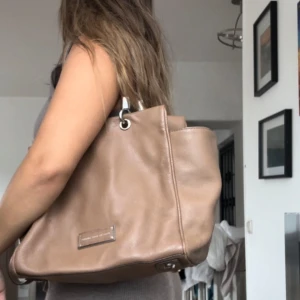 Beige axelväska från Marc by Marc Jacobs - Säljer en snygg beige axelväska från Marc by Marc Jacobs i mjukt skinn. Väskan har silverfärgade metalldetaljer och en diskret logga framtill. Perfekt storlek för att få plats med allt du behöver till vardagen. Den enda skadan  den har är att silver detaljerna har rostat på vissa. Skriv för mer info 