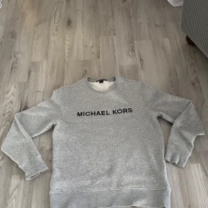 Grå sweatshirt från Michael Kors - Snygg grå sweatshirt från Michael Kors med svart logga framtill. Tröjan har rund halsringning, långa ärmar och är tillverkad i en mjuk bomulls- och polyestermix. Perfekt för en chill och stilren look.