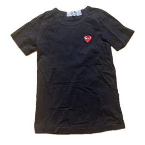 Svart Comme des Garçons Play t-shirt - Svart t-shirt från Comme des Garçons Play med det ikoniska röda hjärtat broderat på bröstet. Klassisk rund halsringning och korta ärmar. Tillverkad i mjuk bomull som känns skön mot huden. Perfekt för en stilren och trendig look. OBS liten i storlek 