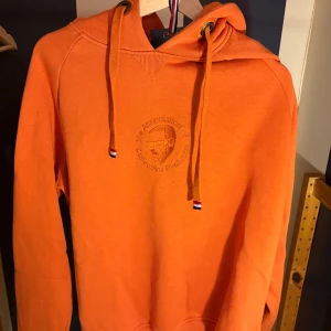 Orange hoodie från WHO's A.C.E - Snygg orange hoodie från WHO's A.C.E med broderad logga på bröstet och coola snören med röd, vit och blå detalj. Tröjan har huva och är tillverkad i mjuk bomull, perfekt för dig som vill sticka ut med färg. Klassisk passform och långärmad.