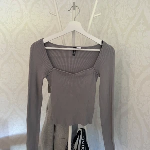 Grå ribbad topp från H&M Divided - Säljer en grå ribbad långärmad topp från H&M Divided. Toppen har en snygg v-ringad skärning och croppad passform. Perfekt att matcha med höga jeans eller kjol för en trendig look.