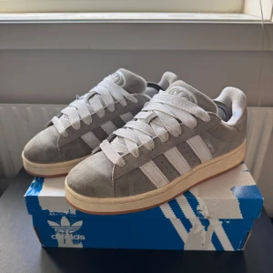 Adidas Campus grå mockasneakers - Adidas Campus sneakers i grå mocka med vita klassiska stripes och vit logga på plösen. Skorna har breda vita snören och en beige mellansula med gummisula undertill. Perfekt för dig som gillar retrovibbar och stilren design.