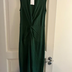 Grön långklänning från MNG - Stilren grön långklänning från MNG med ärmlös design och snygg twistad detalj framtill i midjan. Klänningen har en rak passform och är tillverkad i ett mjukt, lätt material som ger en elegant siluett.