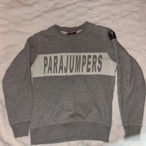 Grå sweatshirt från Parajumpers - Snygg grå sweatshirt från Parajumpers med stort logotryck över bröstet och patch på ena ärmen. Tröjan har rund halsringning, ribbade muddar och är tillverkad i mjuk bomullsblandning. Perfekt för en chill och stilren look.