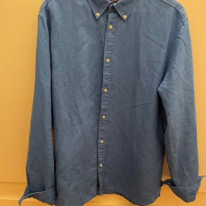 Blå linneskjorta med knappar - Snygg blå skjorta i linne med klassisk krage och vita knappar hela vägen framtill. Skjortan har lång ärm med manschett och en avslappnad passform. Perfekt för dig som gillar stilrena och enkla plagg med en fräsch känsla.