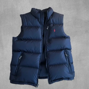 Blå Ralph Lauren väst - Säljer en stilren blå dunväst från Ralph Lauren. Västen är knappt använd och i nyskick. Nypriset för västen ligger runt 2000 kr