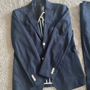 Mörkblå kostym från Riley - Säljer en stilren mörkblå kostym från Riley i storlek 50, slim fit. Kavajen har ljusa knappar och klassisk krage. Kostymen är helfärgad och passar perfekt till formella tillfällen. Byxorna är raka och matchar kavajen. Endast använd 1 gång!!!
