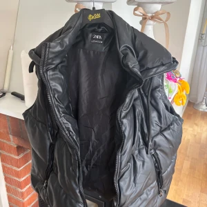 Svart vadderad väst från Zara - Säljer en svart vadderad väst från Zara med hög krage och dragkedja framtill. Västen har två fickor med dragkedja och en quiltad design som ger en modern look. Perfekt att slänga över en hoodie eller tröja.