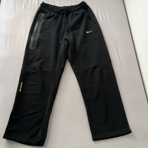 Svarta NOCTA x Nike mjukisbyxor - Svarta mjukisbyxor från Nike och NOCTA med gul logga och gul swoosh. Byxorna har dragsko i midjan, raka ben och en lång dragkedjeficka på höger sida. Perfekta för en sportig och avslappnad stil. 