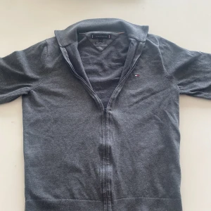Grå ziptröja från Tommy Hilfiger - Säljer en stilren grå ziptröja från Tommy Hilfiger i mjuk bomull. Tröjan har lång ärm, klassisk krage och dragkedja framtill. Liten broderad logga på bröstet. Perfekt för en avslappnad och snygg look. Liten i storlek