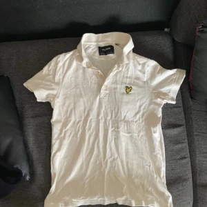 Vit piké från Lyle & Scott - Vit pikétröja från Lyle & Scott med klassisk krage och knappar framtill. Tröjan har det ikoniska broderade gula örnloggan på bröstet. Tillverkad i 100% bomull för en skön och luftig känsla.