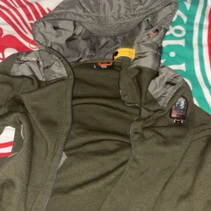 Grön hybridjacka från Parajumpers - Säljer en grön hybridjacka från Parajumpers med grå huva och detaljer. Jackan har dragkedja framtill, lång ärm och klassisk Parajumpers-logga på ärmen. Perfekt för övergångssäsonger och har en sportig look.