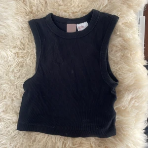 Svart ribbad linnetopp från H&M - Säljer en svart ribbad linnetopp från H&M. Toppen är ärmlös och har en croppad passform med breda axelband och rund halsringning. Perfekt att styla till jeans eller kjol.