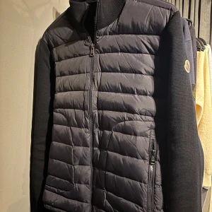  Moncler cardigan, blå - Snygg blå cardigan från Moncler med quiltad front och ärmar i stickad bomull. Jackan har dragkedja framtill, ståkrage och Moncler-logga på ärmen. Perfekt för dig som vill ha en stilren och exklusiv look. Jackan är i storlek M och jag säljer den då den börjar bli lite kort, den är näst intill ny och har inga defekter. Tveka inte om att höra av dig om du har frågor.