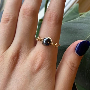 Guldfärgad ring med svart sten ”hematit” - Säljer en snygg guldfärgad ring med en rund svart sten i mitten. Ringen har en unik tråddesign runt stenen som ger ett handgjort intryck. Perfekt accessoar för att lyfta din stil.