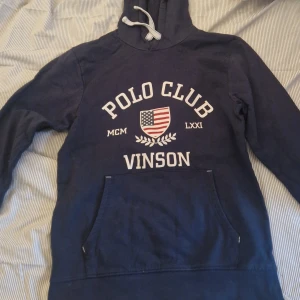 Mörkblå hoodie från Vinson Polo Club - Säljer en mörkblå hoodie från Vinson Polo Club med vit text och ett amerikanskt flaggmärke på bröstet. Tröjan har huva med dragsko och en stor magficka framtill. Perfekt för en avslappnad stil.