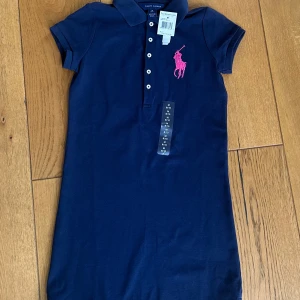 Marinblå pikéklänning från Ralph Lauren - Säljer en klassisk marinblå pikéklänning från Ralph Lauren med rosa broderad logga på bröstet. Klänningen har korta ärmar, krage och knappar framtill. Aldrig använd, lappar sitter kvar!! Storlek barn M eller 8-10 år.