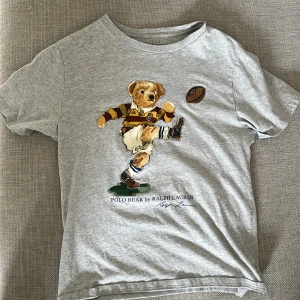 Grå t-shirt med Polo Bear från Ralph Lauren - Säljer en grå t-shirt från Ralph Lauren med den ikoniska Polo Bear som spelar rugby på bröstet. T-shirten har rund hals och korta ärmar. Perfekt för dig som gillar sportiga och klassiska motiv.