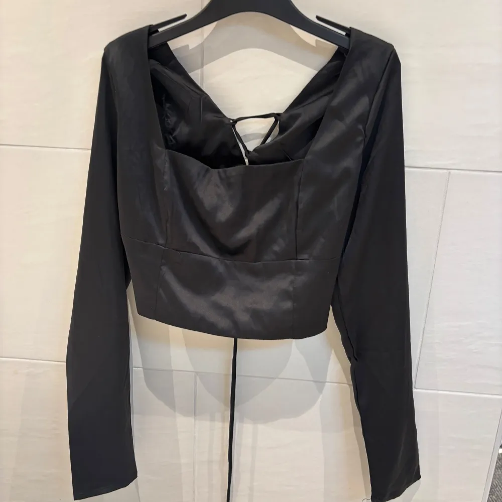 Snygg svart croppad topp i satinliknande material med långa ärmar och unik snörningsdetalj i ryggen. Toppen har en djup urringning och sitter figurnära, perfekt för att skapa en trendig look. Helt oanvänd med etikett på. Yläosat.