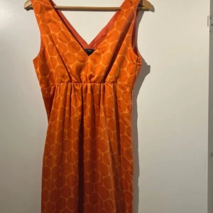 Orange ärmlös klänning från H&M - Säljer en orange ärmlös klänning från H&M med v-ringning och diskret cirkelmönster. Klänningen har dragkedja i sidan och en lätt utställd passform. Perfekt för sommarens tillfällen!