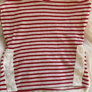 Randig stickad tröja från Zara - Säljer en vit och röd stickad tröja från Zara med horisontella ränder. Tröjan har lång ärm och dekorativa vita knappar på sidorna. Perfekt för dig som gillar klassiska mönster med en twist!