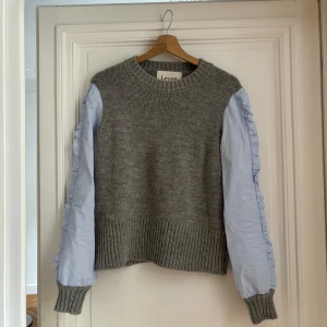 Grå stickad tröja med ljsblå ärmar från Levate - Säljer en unik grå stickad tröja från Levate med ljusblå, volangprydda ärmar i kontrastmaterial. Tröjan har rund halsringning och ribbade muddar. Så unik och perfekt till våren/sommar!!