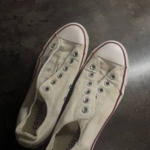 Vita Converse All Star sneakers - Klassiska vita Converse All Star sneakers med röd och blå rand längs sulan. Lågt skaft och rund tå, tillverkade i canvas med gummisula. Perfekta för en avslappnad och tidlös stil. Inga skosnören