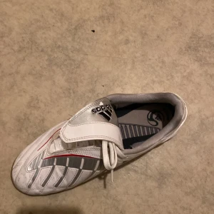 Adidas vita fotbollsskor med svarta och röda detaljer - Säljer ett par vita Adidas fotbollsskor med svarta ränder och röda detaljer. Skorna har snörning och en täckande rem över snörningen samt mönstrad sula för inomhusspel. Klassisk design med rund tå och syntetmaterial.