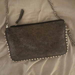 Svart axelväska med nitar från Zara Woman - Säljer en svart axelväska från Zara Woman med silverfärgade nitar längs kanterna och kedjerem. Väskan har dragkedja upptill och flera fack inuti. Perfekt för att bära det viktigaste med stil.