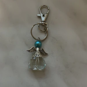Nyckelring med ängel i glas och silverdetaljer - Söt nyckelring med en ängel gjord av klar glasdroppe, silverfärgade vingar och en blå pärla som huvud. Perfekt som accessoar till väskan eller nycklarna.