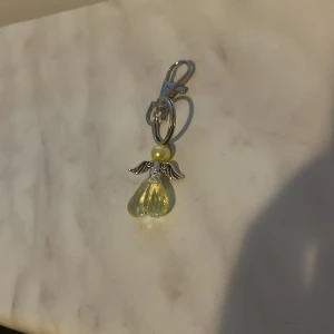 Ängelberlock med pärla och vingar - Söt accessoar i form av en ängel med silverfärgade vingar, pärldetalj och en droppformad gul transparent sten. Perfekt att hänga på väska eller nyckelring.