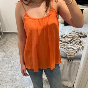 Orange topp med knytband - Säljer en luftig orange topp med smala axelband som knyts upptill. Toppen har en lös passform och är tillverkad i ett mjukt, lätt tyg som passar perfekt för varma dagar. Mycket bra skick!