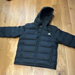 Svart dunjacka med huva från Adidas - Säljer en svart dunjacka från Adidas med huva och dragkedja framtill. Jackan har två sidofickor med dragkedja och klassisk Adidas-logga på bröstet. Perfekt för kyliga dagar. Storlek L men passar M. Säljs för 300