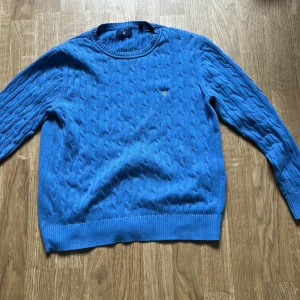 Blå kabelstickad tröja från Gant - Säljer en blå kabelstickad tröja från Gant med rund halsringning och diskret logga på bröstet. Tröjan har långa ärmar och ribbade muddar vid ärmslut och nederkant. Perfekt för lager-på-lager och snygg till jeans.