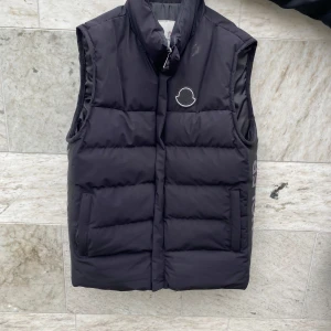 Svart dunväst från Moncler - Säljer en svart dunväst från Moncler med broderad logotyp på bröstet. Västen har två fickor framtill och stängs med dragkedja. Klassisk quiltad design som passar perfekt till lager-på-lager-stil.