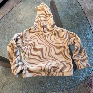 Beige och vit fleecehoodie från Lindex - Mysig hoodie i fleece från Lindex med beige och vitt vågigt mönster. Tröjan har huva och långa ärmar, perfekt för att hålla sig varm och bekväm. Supermjuk och fluffig känsla!