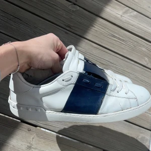 Valentino Garavani sneakers i vitt och blått - Snygga Valentino Garavani sneakers i vitt med en bred blå rem över snörningen. Skorna har en rund tå och är tillverkade i skinn. Perfekta för en stilren look. Skorna är väldigt slitna på vissa ställen och därav har jag dragit av priset sjukt mycket 🌟💪