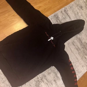 Svart hoodie med tryck från Trapstar - Säljer en svart hoodie från Trapstar med stort grafiskt tryck på ryggen och text på bröstet. Ärmarna har röda tryck längs hela sidan. Klassisk känguruficka och huva med dragsko. Perfekt för dig som gillar streetwear. Gjort efter collaben på J hus album ' Beautiful and brutal yard ' 