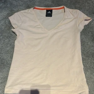Ljusbeige t-shirt från Adidas - Ljusbeige t-shirt från Adidas med v-ringning och korta ärmar. T-shirten har en diskret orange kant på insidan av halsen och är tillverkad i mjuk bomull. Perfekt för en sportig och avslappnad stil.